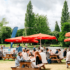 PEOPLE SITTING AT PICNIC TBALES WITH UMBRELLAS M SITUATED IN FRONT OF THE RIVER AT HENLEY ROYAL REGATTA