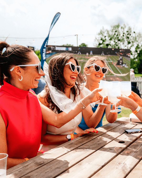 3 WOMEN IN WHITE HEART SHAPED SUNGLASSES CHERS OVER A PICNIC TABLE OUTSIDE IN THE SUNSHINE