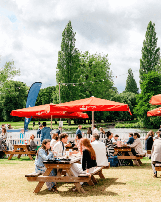 PEOPLE SITTING AT PICNIC TBALES WITH UMBRELLAS M SITUATED IN FRONT OF THE RIVER AT HENLEY ROYAL REGATTA