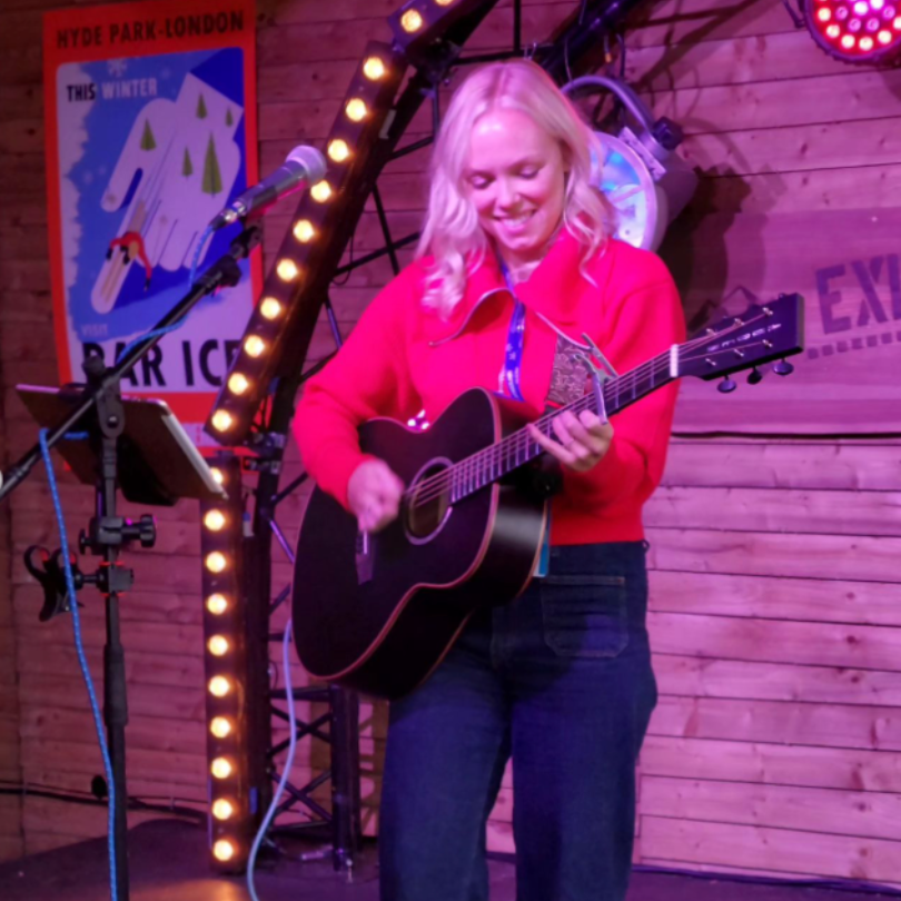 female singer wearing a red jacket sings on a stage with her guitar