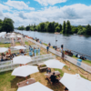 A sunny birdseye view of the Pavilions Enclosure, showing white pavilions with white picket fences alongside the River Thames during Henley Royal Regatta