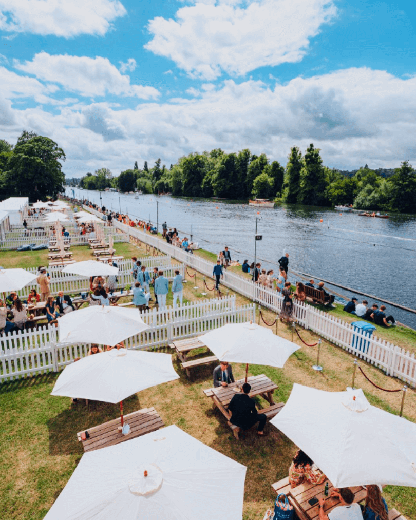 A sunny birdseye view of the Pavilions Enclosure, showing white pavilions with white picket fences alongside the River Thames during Henley Royal Regatta