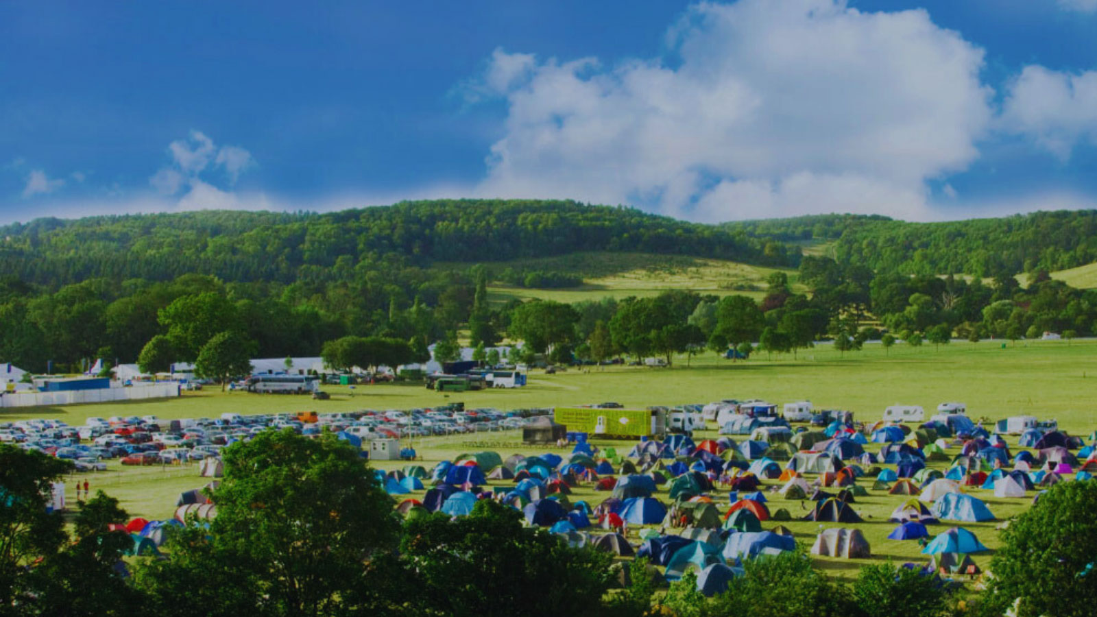 Viewof a campsite set on green fields with lush green hills behind. The sky is blue and the sun is shining