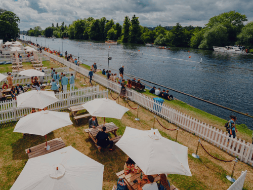 Multiple picnic benches and umbrellas with people sitting at them by the side of a river