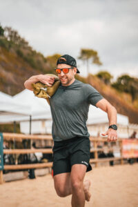male in grey tee shirt and black shorts carries a heavy weight while working out on beach