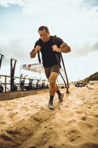 Man in black workout gear runs along beach with two heavy ropes