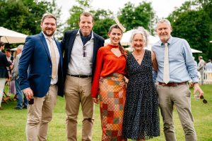 a group of mid aged men and women in shirts, ties and summer dresses stand to have their photo taken at an outdoor event