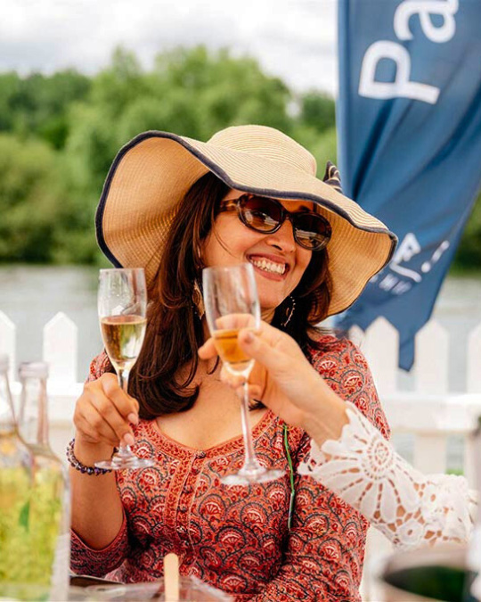 A woman wearing a hat holding a glass of champagne and smiling at someone outside of the frame