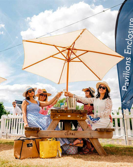 4 Women drseed up for an event sit at a picnic table under an umbrella and cheers with their drinks