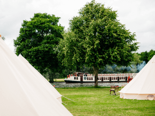 Two cream-coloured bell tents set up on grass near a river, with trees in the background and a boat passing on the water.