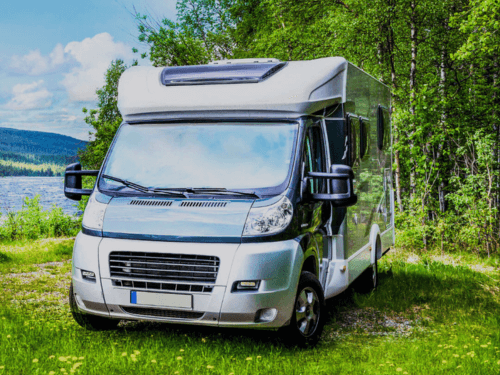 A grey motorhome parked on grass beside a river, surrounded by trees and greenery.