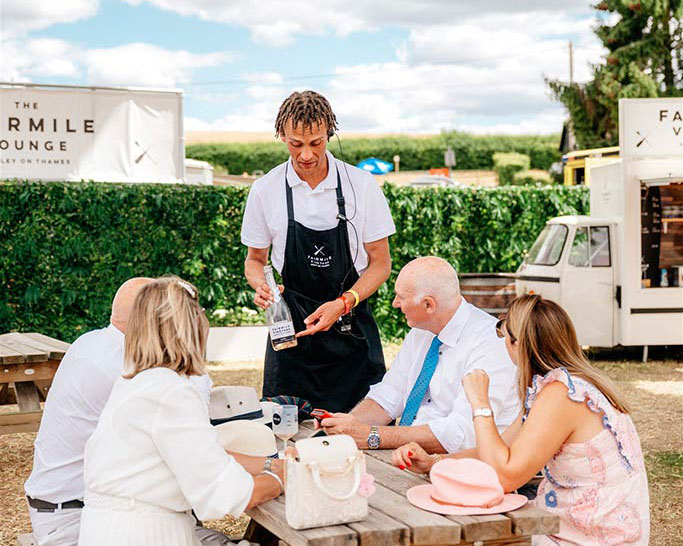 4 people of varying ages sit at a picnic ben while a waiter shows them a bottle of wine