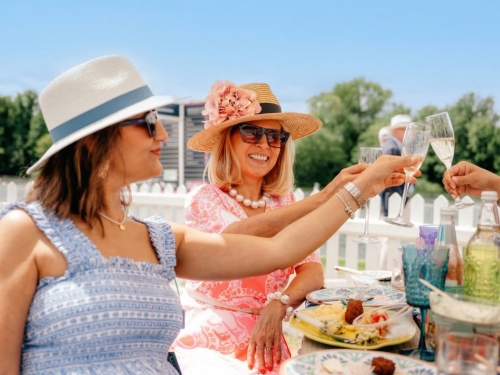 2 older women in hats and dresses clink champagne glasses together at an outdoor event